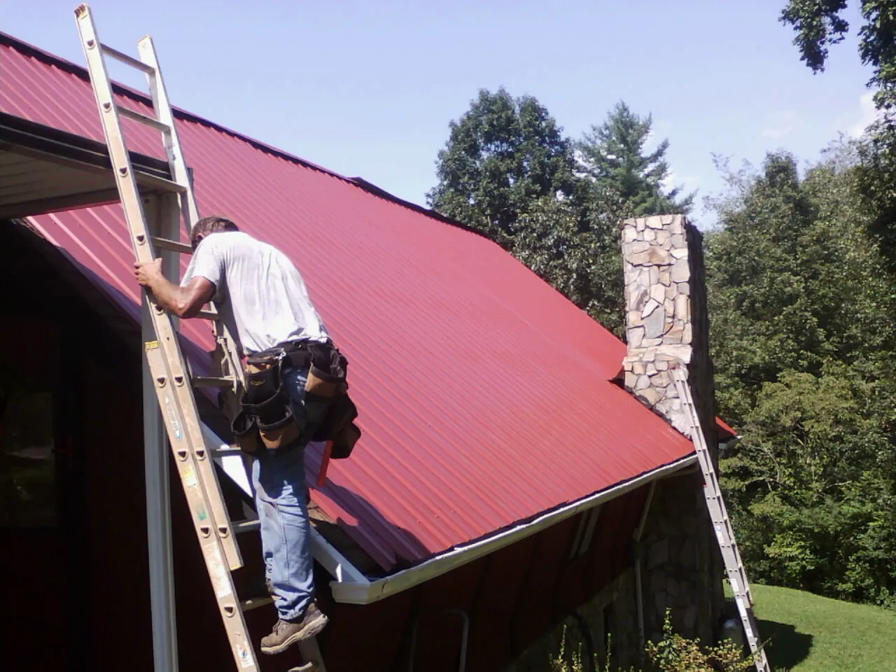 Roofer on a ladder beside a red metal roof and stone chimney — Soffit & Fascia Repair by Roofing Brothers in Sweet Home